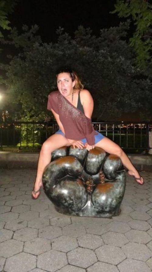 Woman playfully straddles giant fist sculpture, night shot